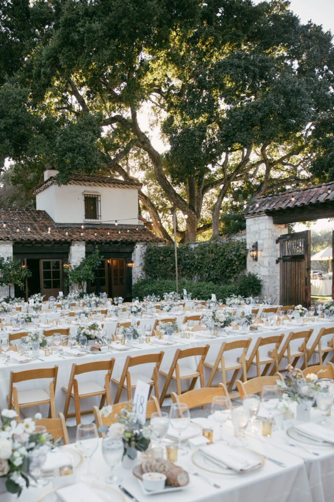 Outdoor reception setup at Holman Ranch with wooden chairs, long tables adorned with floral centerpieces, and string lights under oak trees.