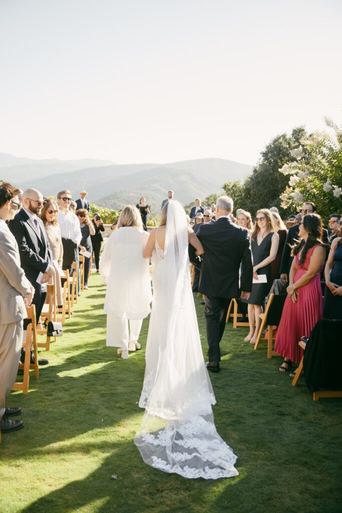 Bride walking down the aisle at Holman Ranch with mountains in the background, surrounded by guests seated on wooden chairs.