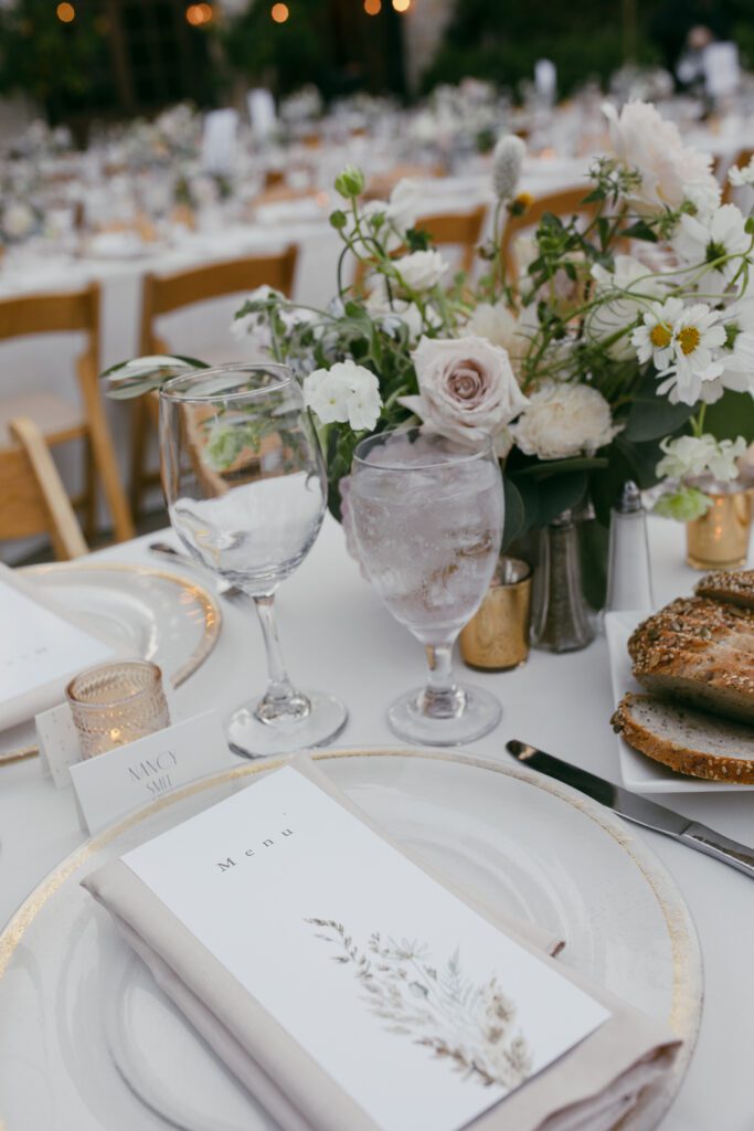 Close-up of a beautifully set table with a floral centerpiece, menu, wine glasses, and candlelit accents at the Holman Ranch wedding reception.